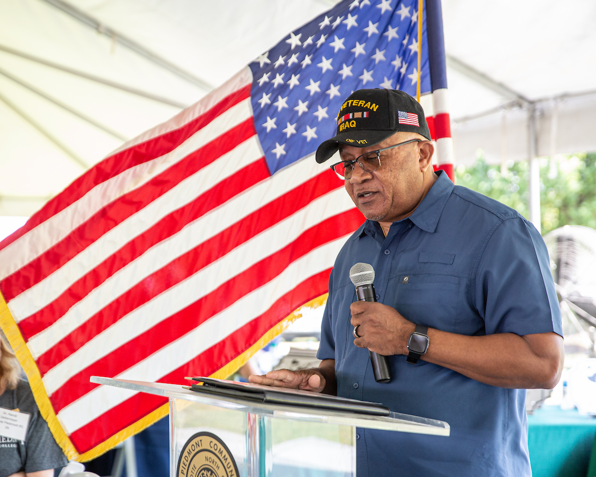 US veteran speaking into microphone at lectern with large American flag waving closely in background.