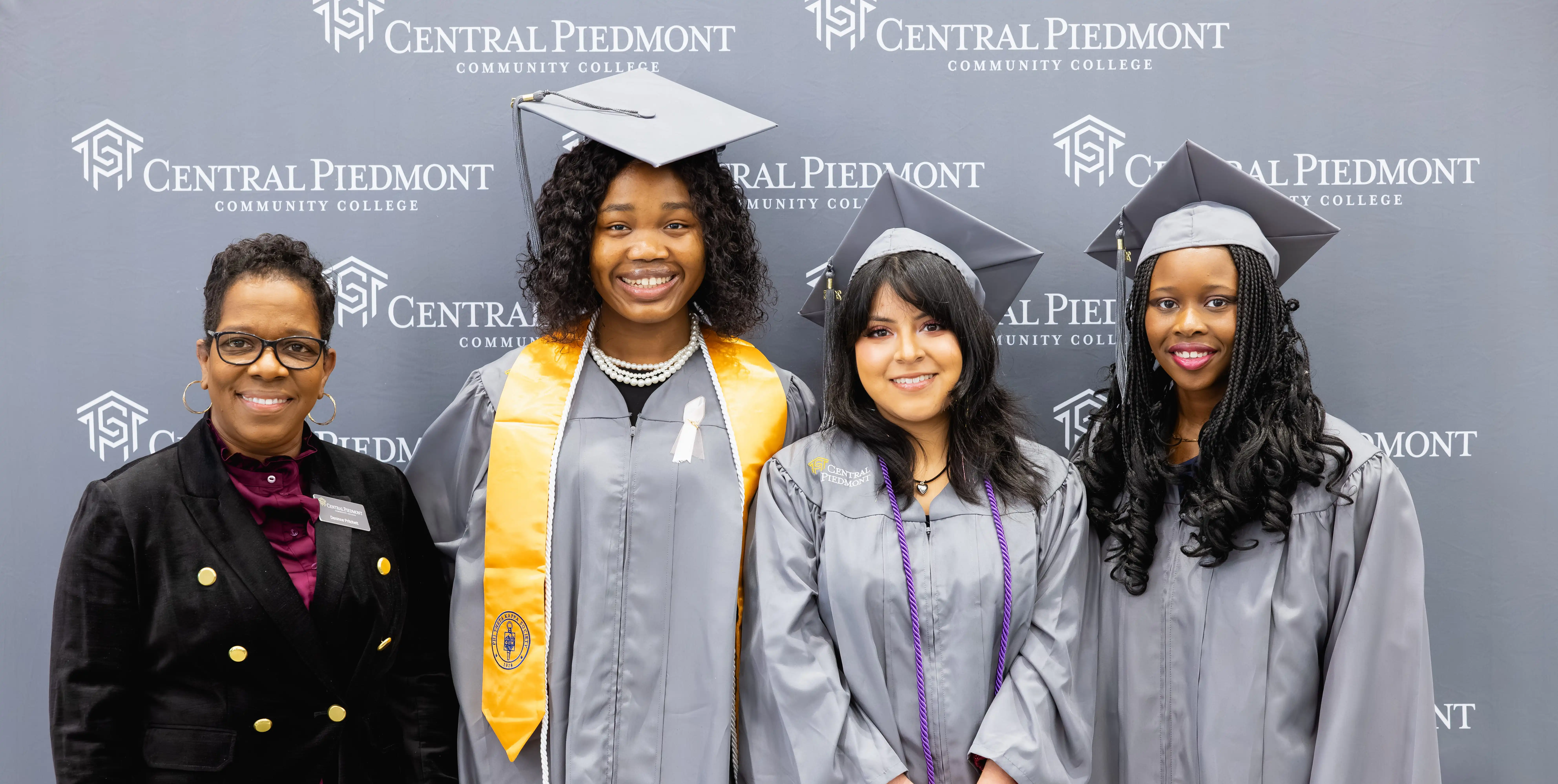 Graduates posing with a staff member in front of a Central Piedmont backdrop