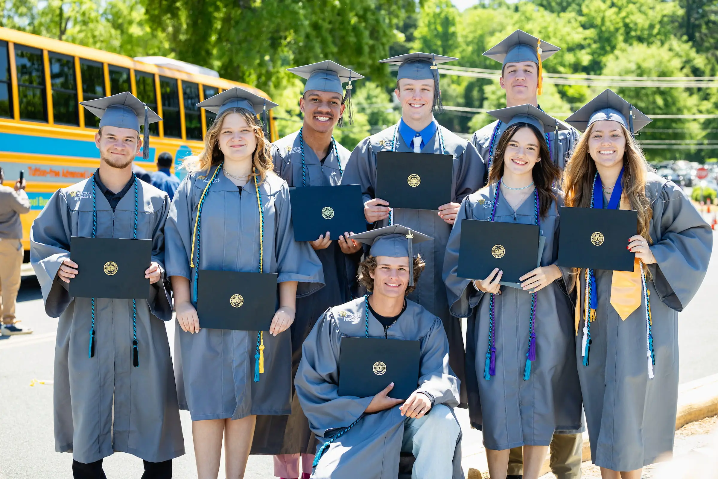 High school graduates in caps and gowns holding diplomas beside a school bus
