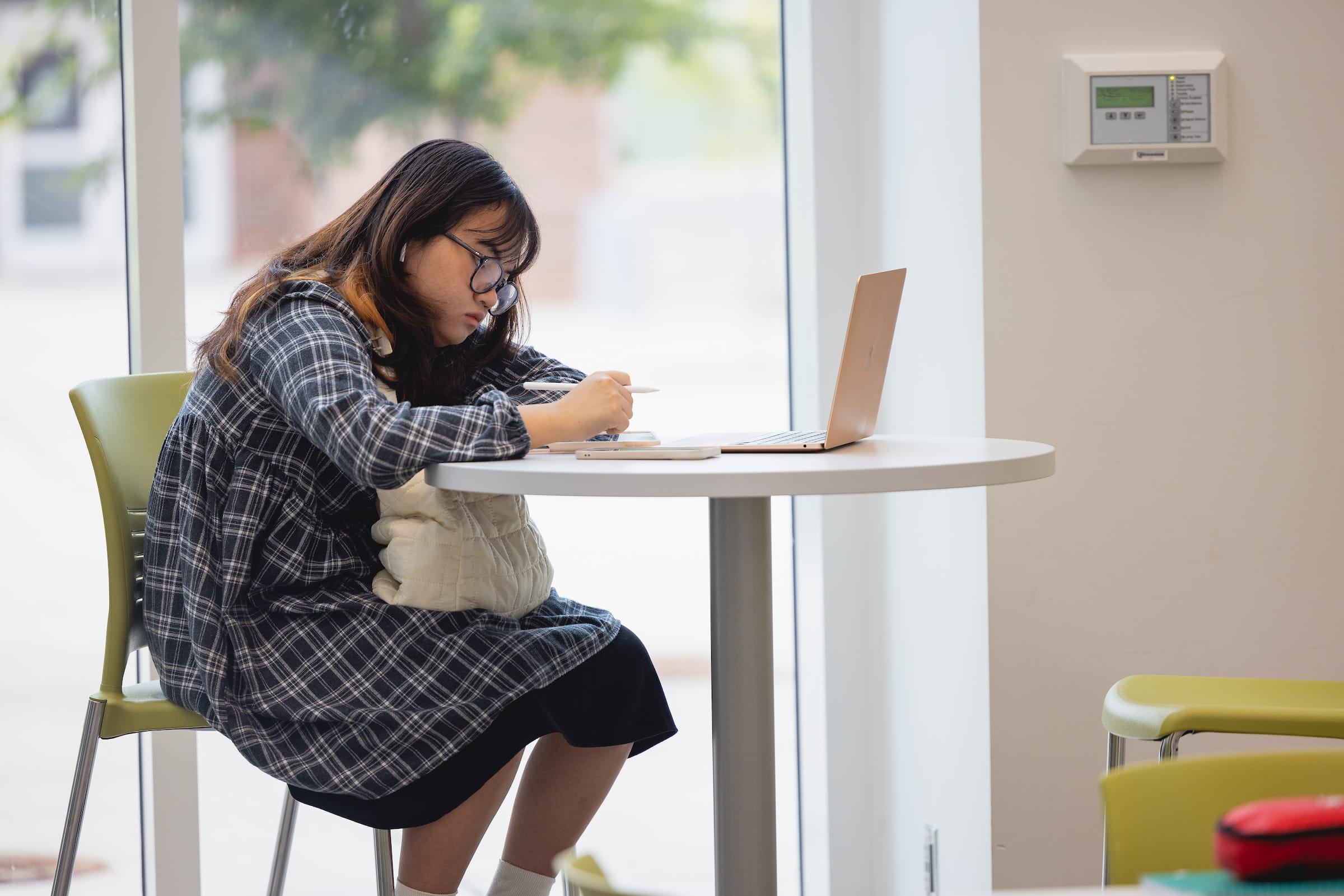 Central Piedmont student concentrating hard on notes at small desk in airy common area with laptop open.