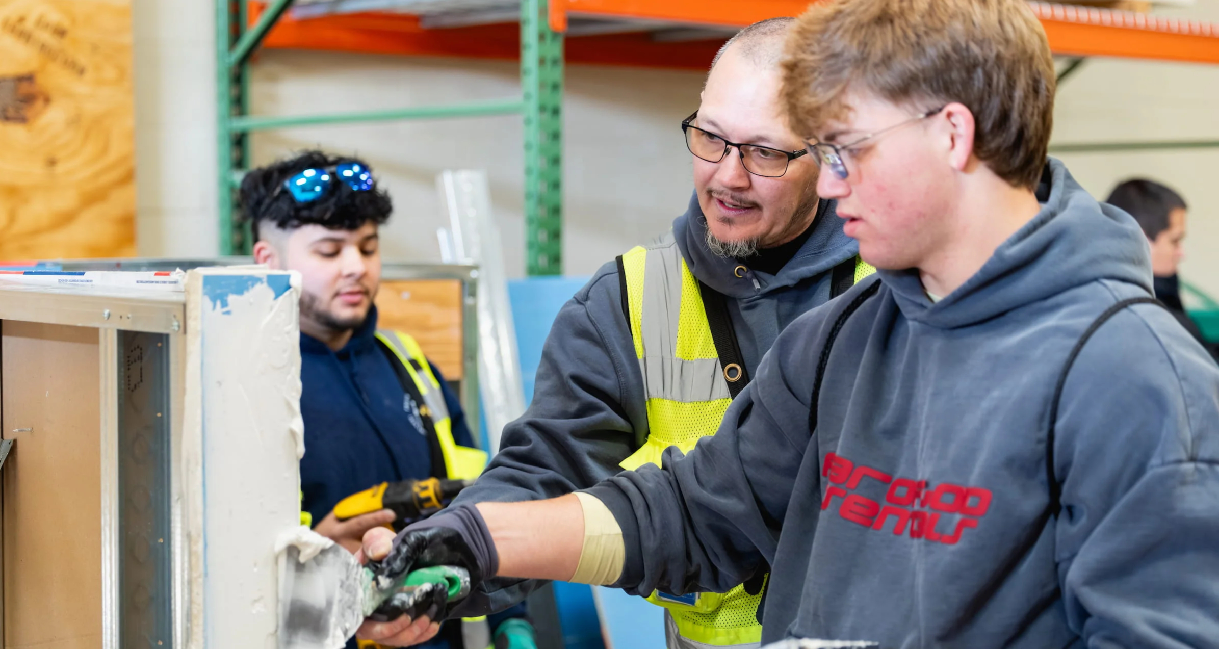 Instructor in a high-visibility vest guiding an apprentice with a tool