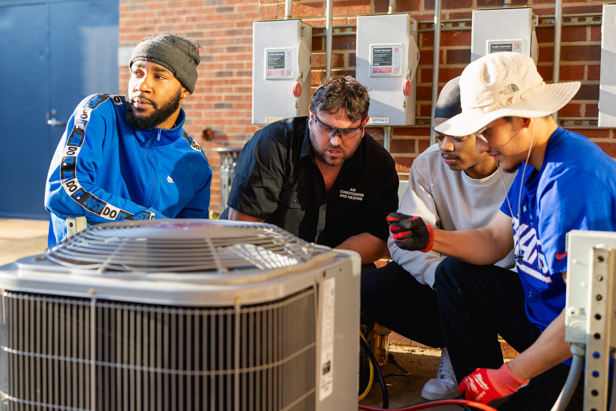 Three Central Piedmont students and instructor kneeling and looking at HVAC unit outside brick building.