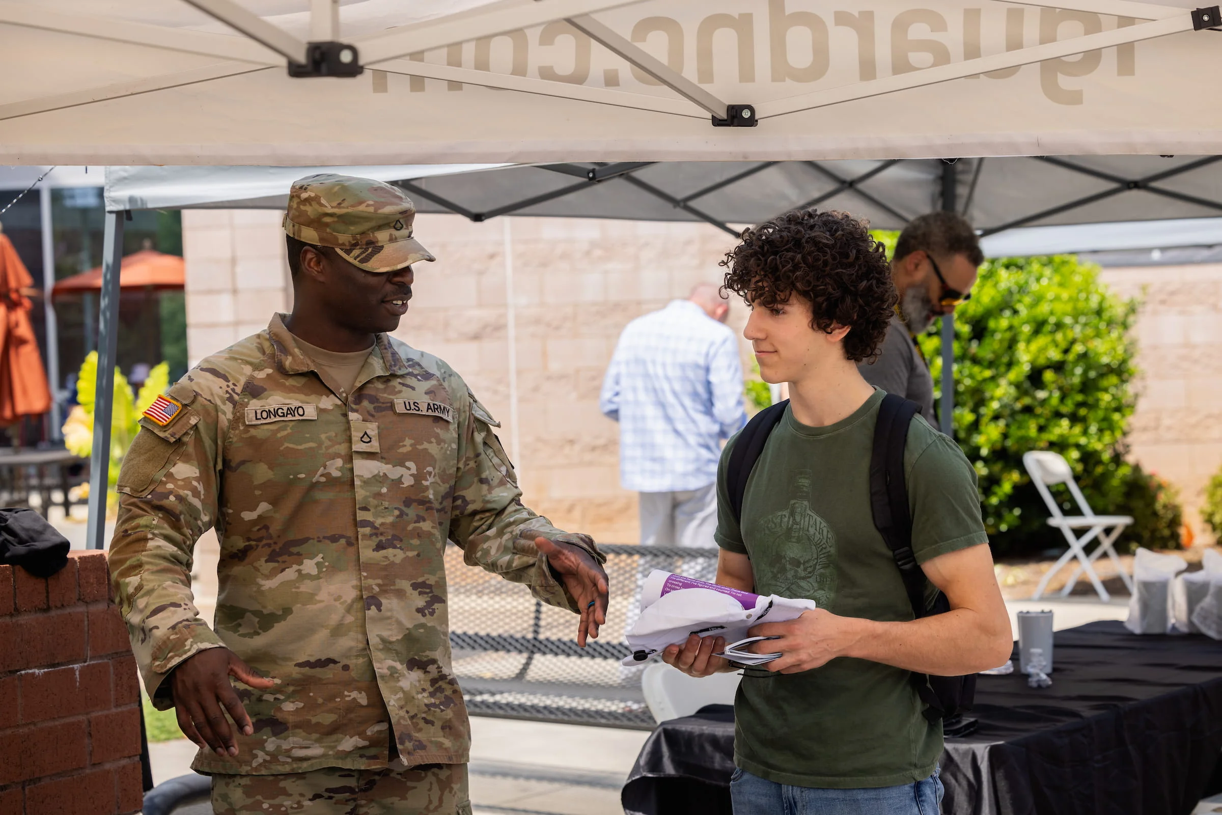 Service member in uniform talking with a student at an outdoor information tent