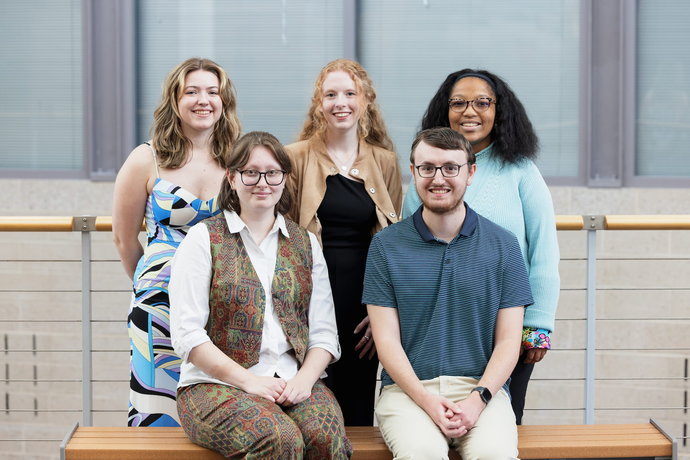 Five students posing together on a bench outside a campus building