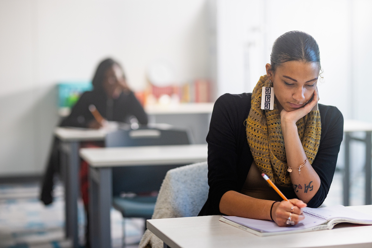 Central Piedmont student concentrating hard looking down at notebook in classroom with another student in background.