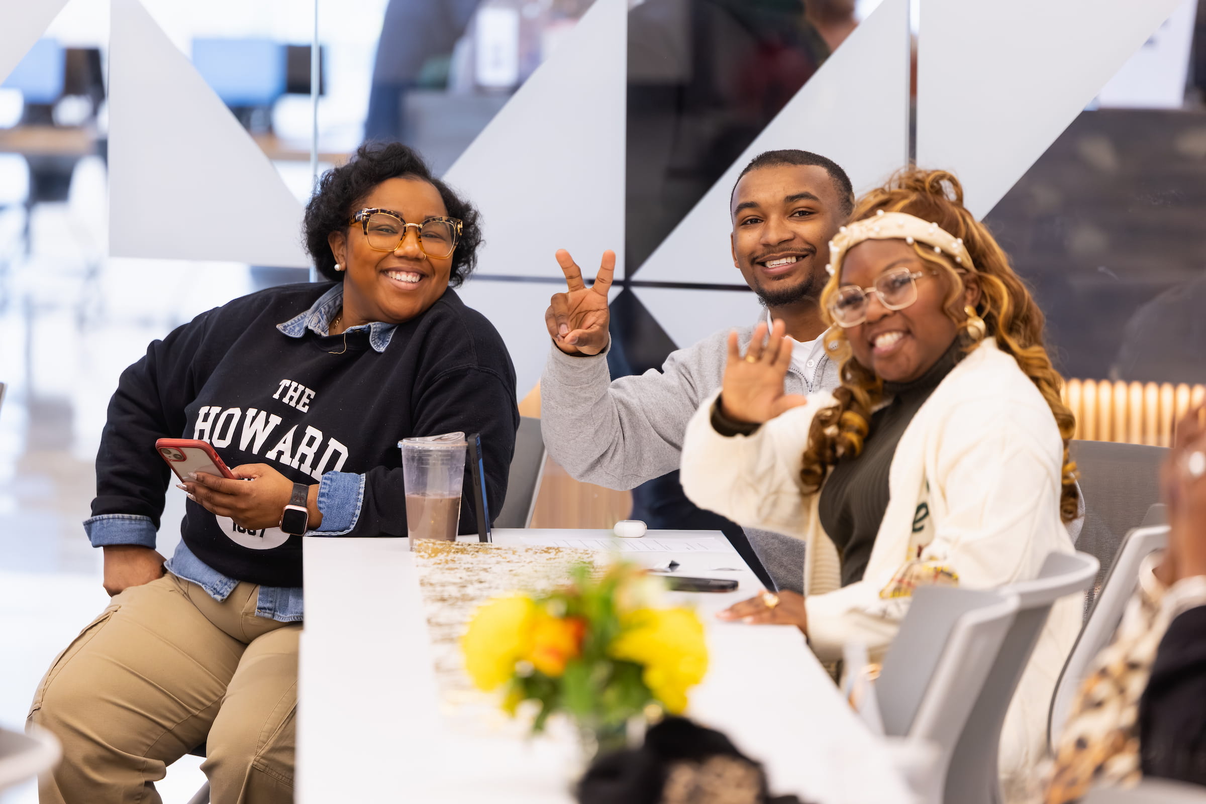 Three Central Piedmont students sitting at end of long white table inside, smiling and waving at camera.