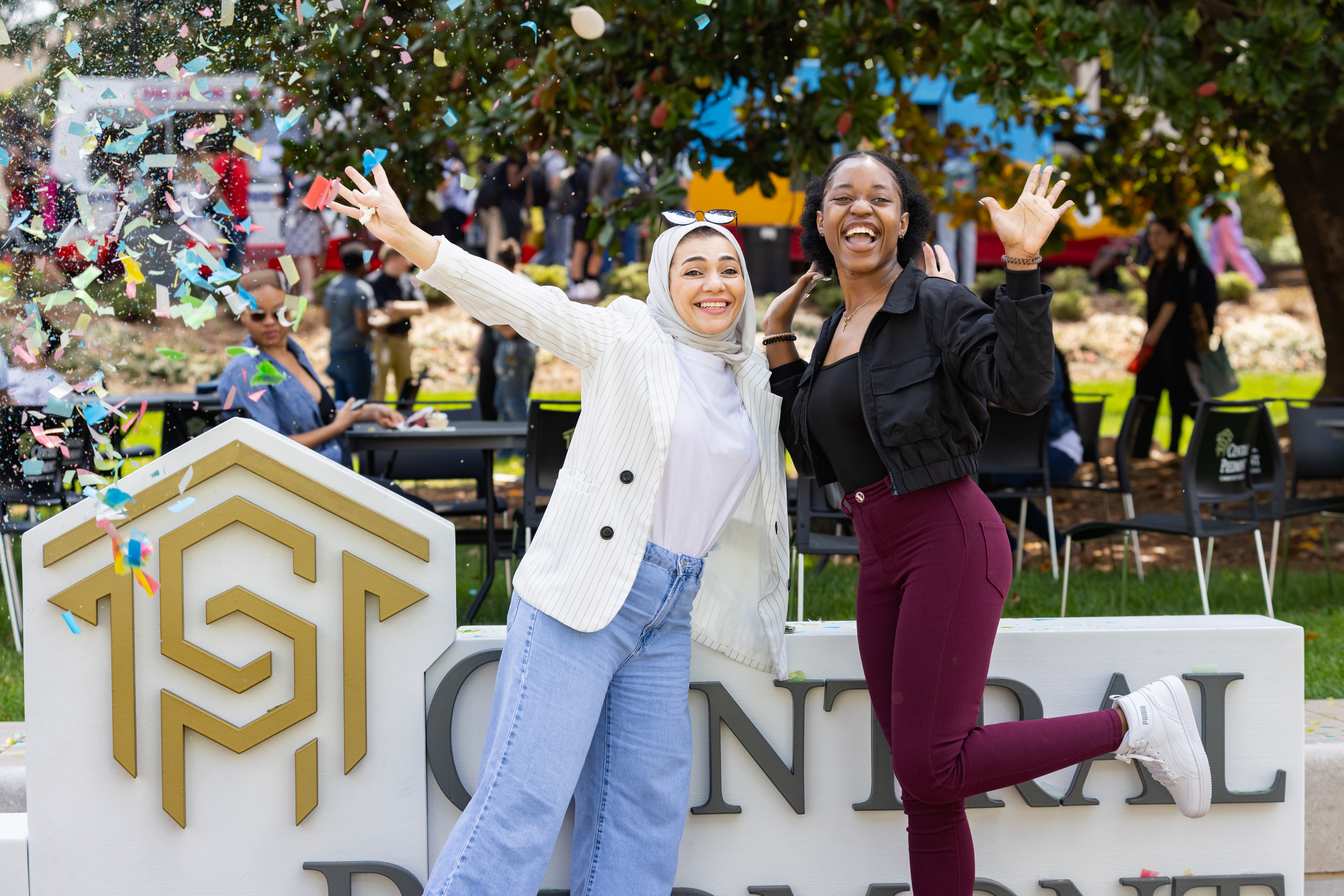 Two students celebrating and posing for camera in front of large Central Piedmont signage with confetti all around.
