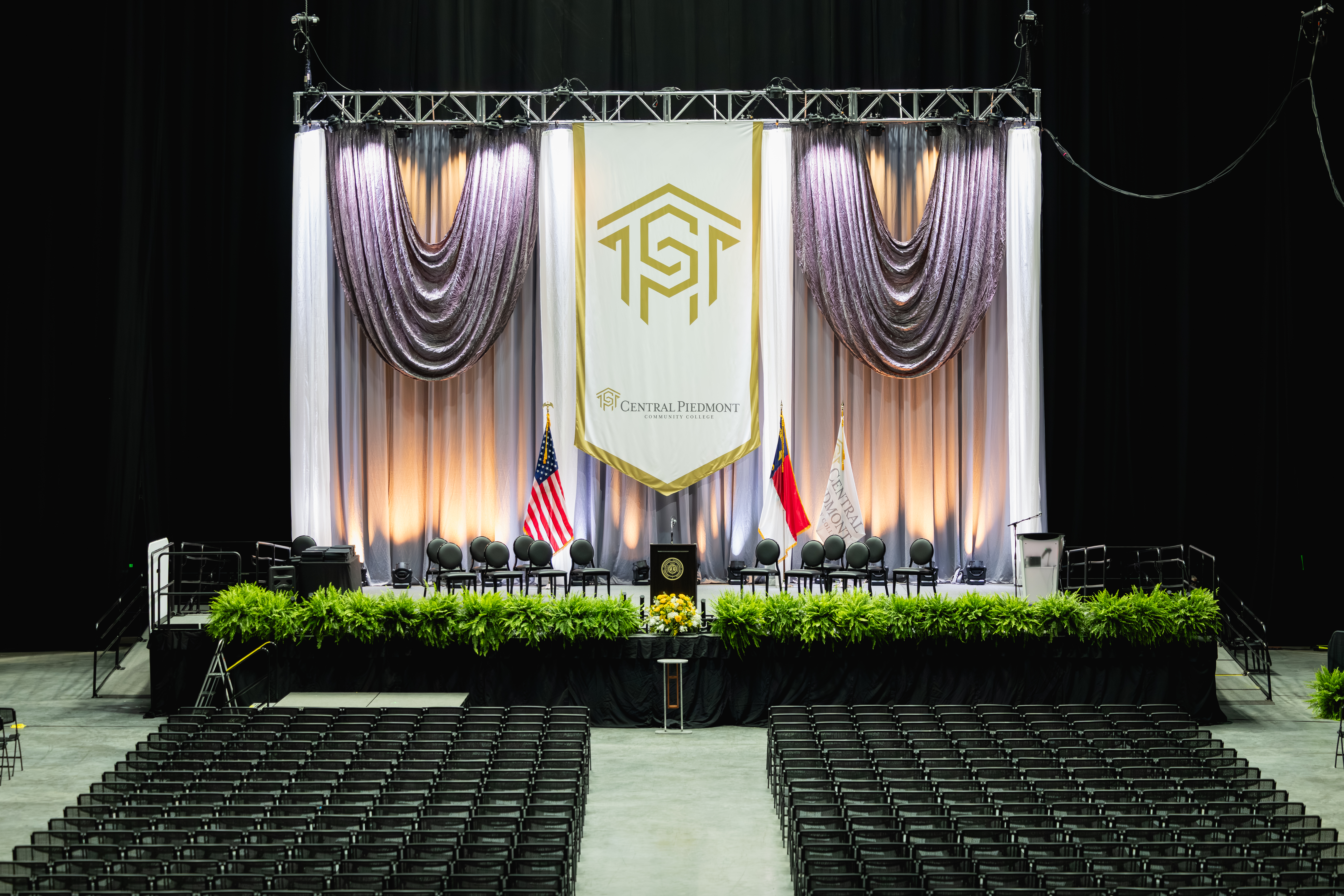 Graduation ceremony stage with draped curtains, greenery, flags and chairs
