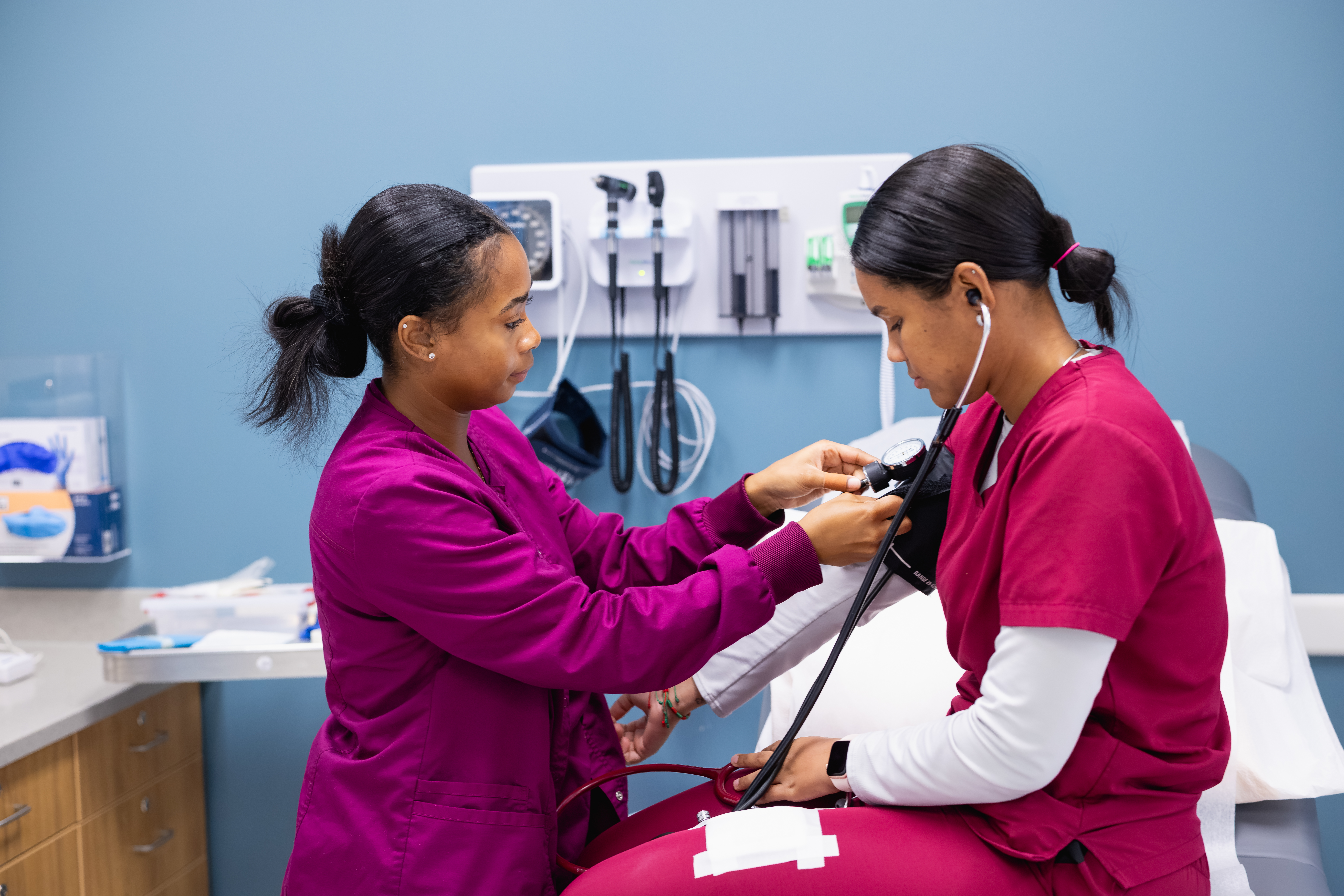 Medical student taking blood pressure of another student who is sitting on an exam table.