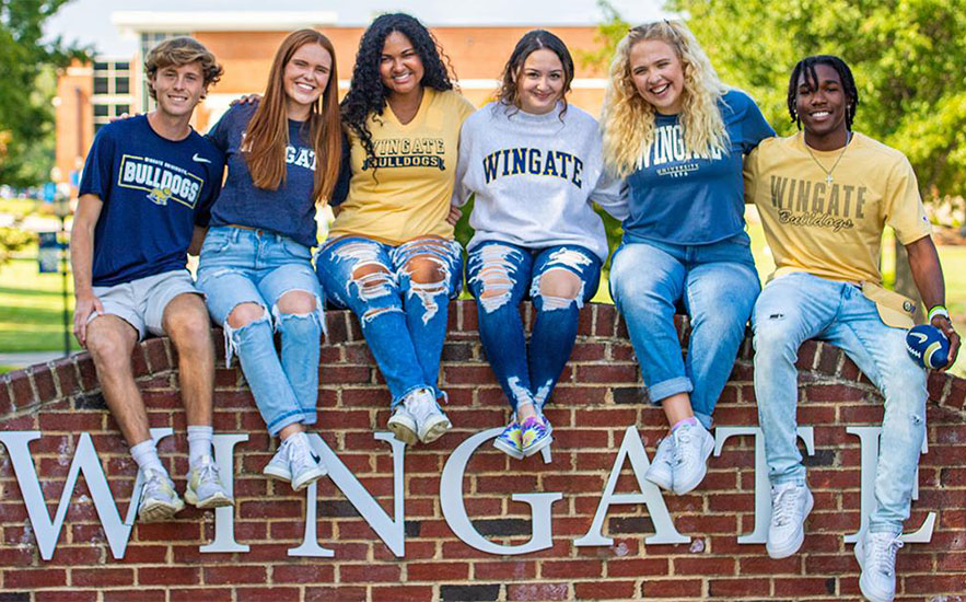 Students in Wingate shirts sitting on the university's brick entrance sign