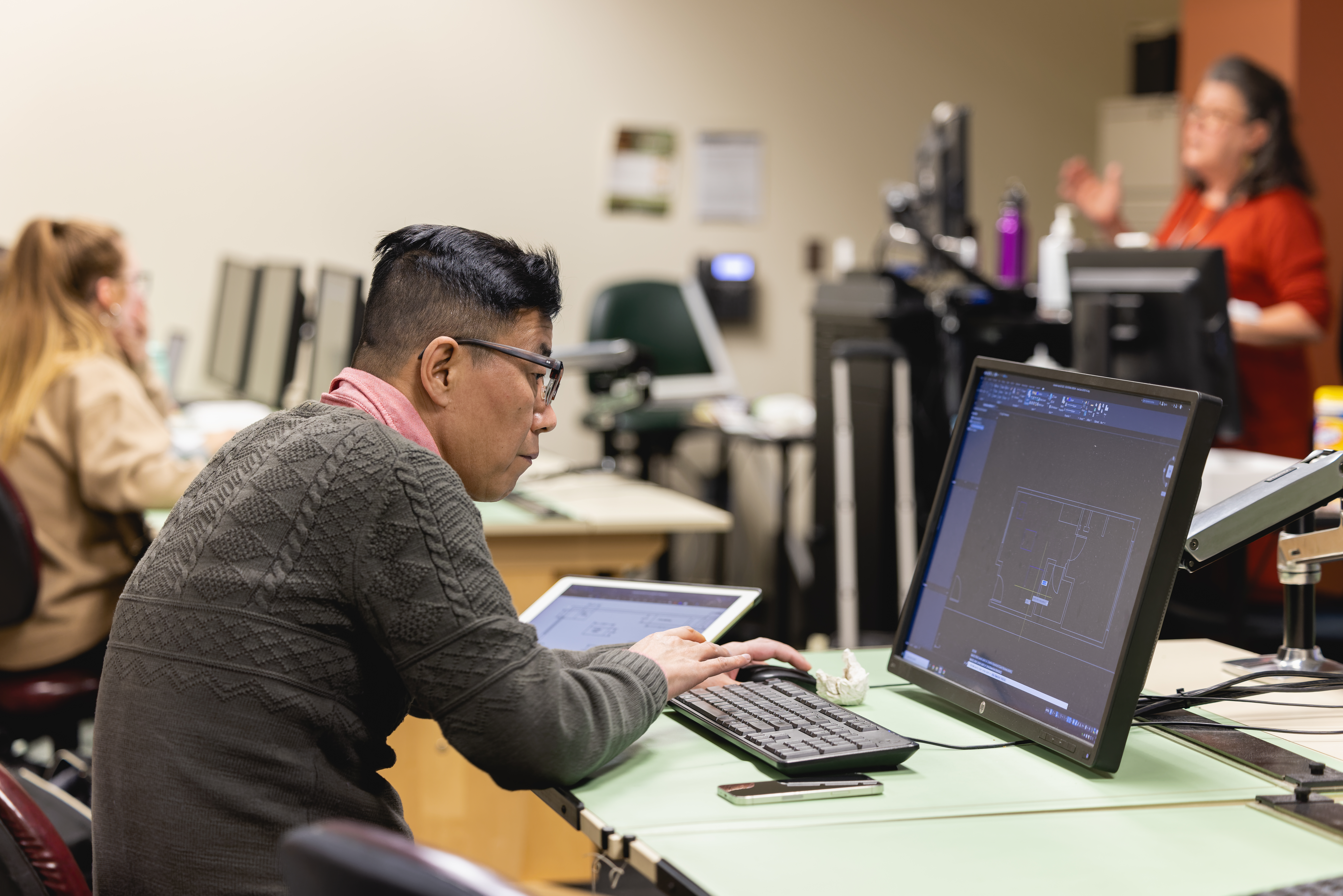 Student looking intently at a computer screen with another student and instructor in the background