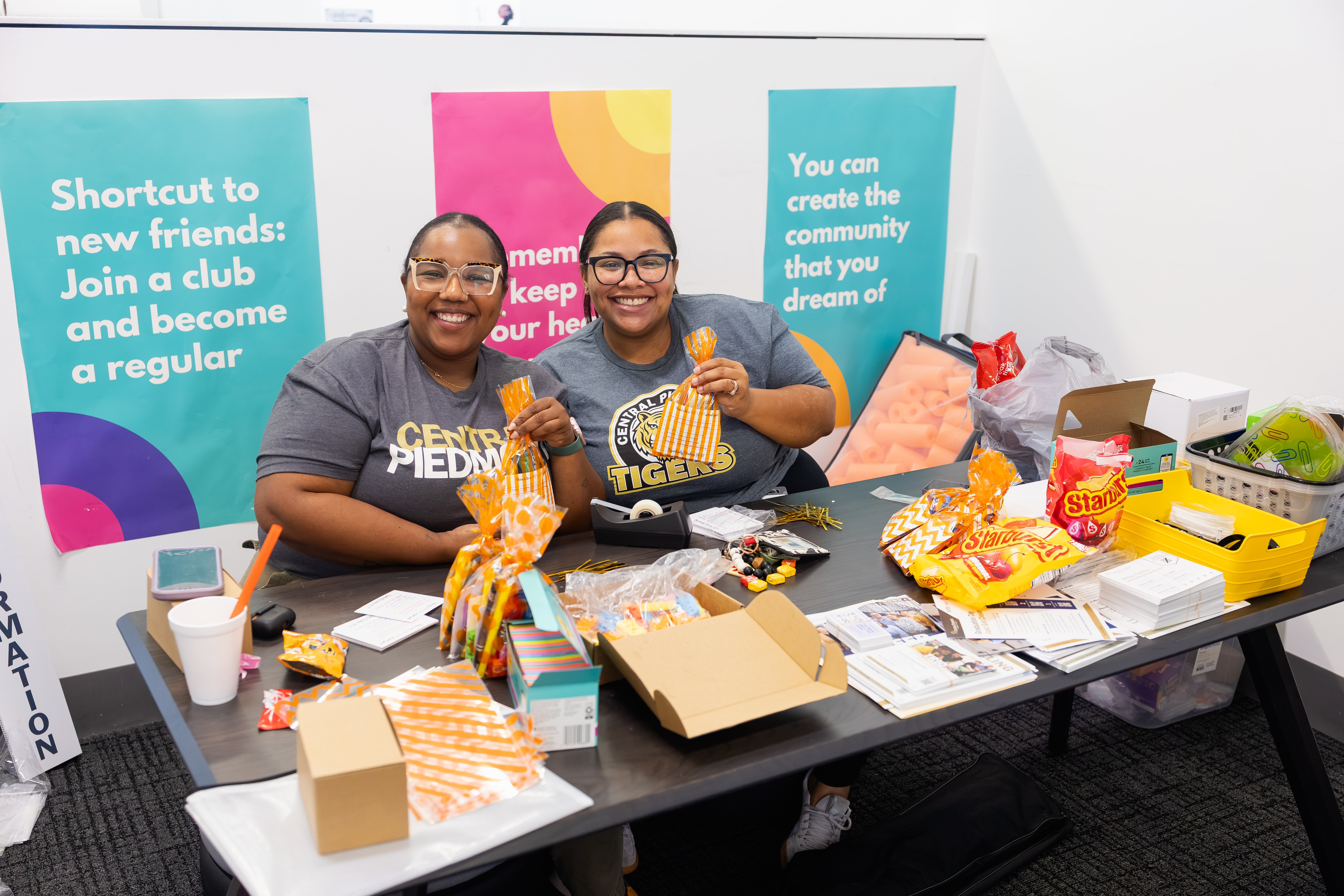 Two staff members wearing Central Piedmont shirts behind a table with papers, snacks and gift bags. Behind them are banners saying "Join a club" and "You can create the community that you dream of".