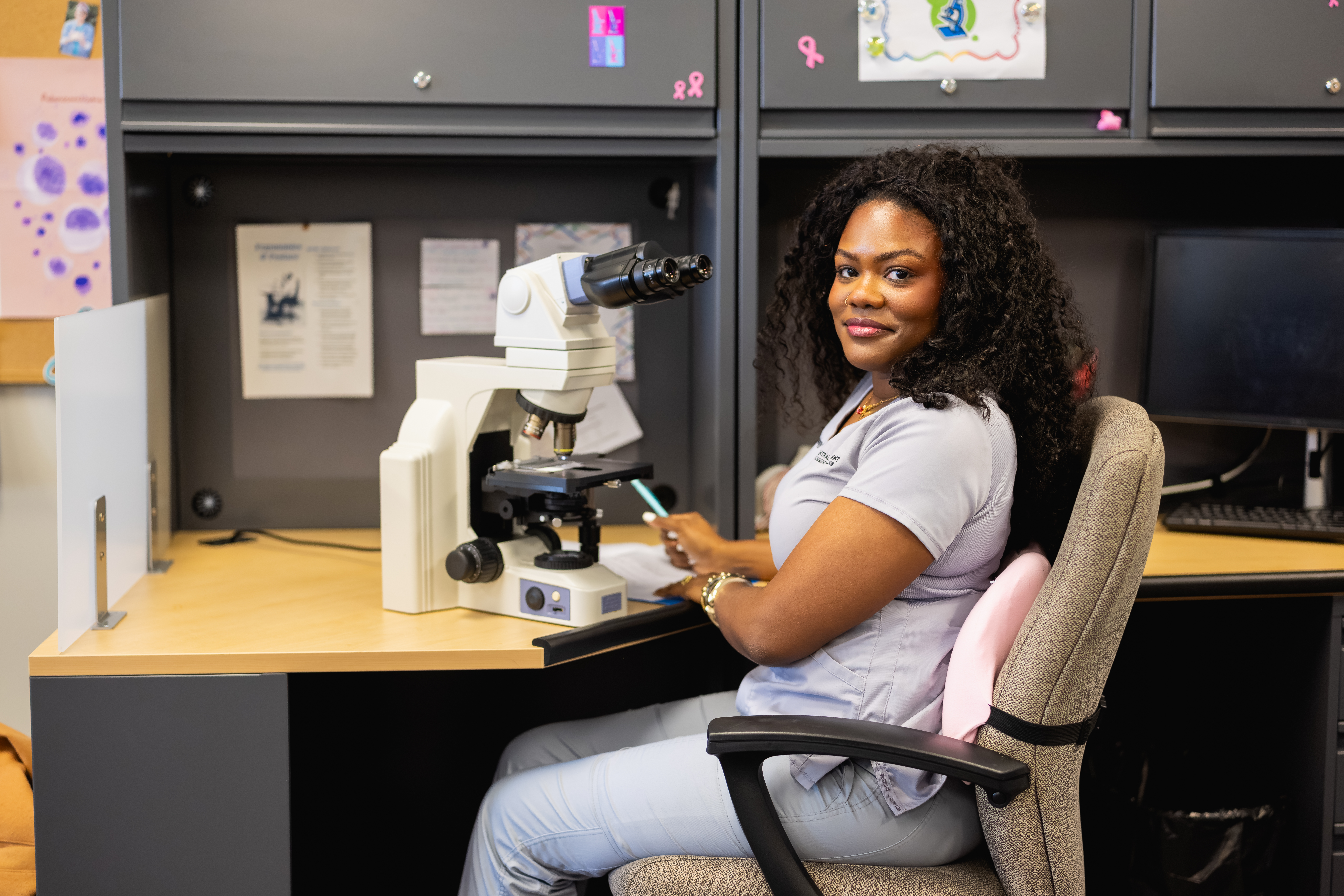 Student at a desk wearing medical scrubs and sitting behind a microscope turned toward the camera and smiling
