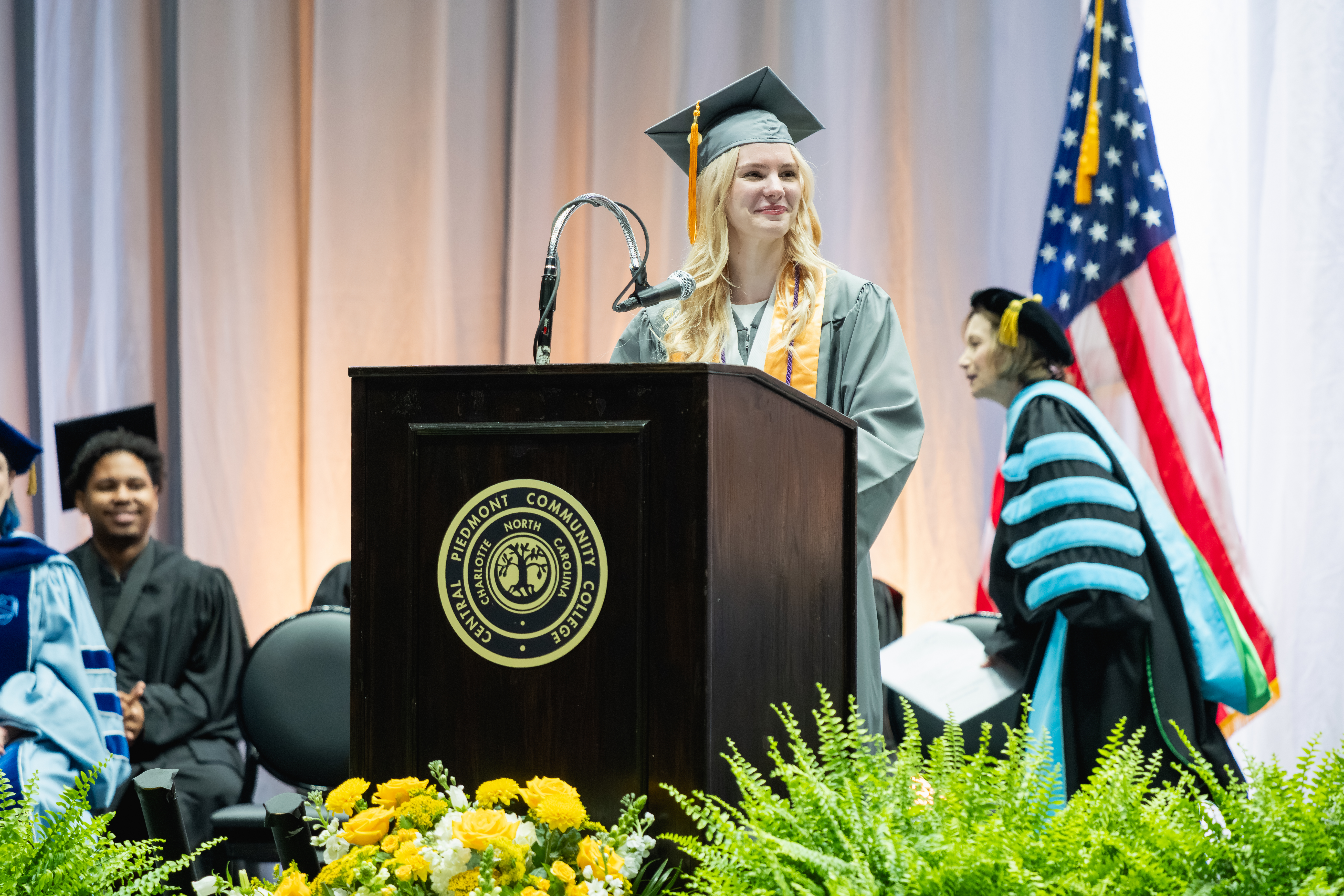 A graduate in academic regalia stands at a podium with the emblem of Piedmont Community College, addressing an audience during a commencement ceremony.