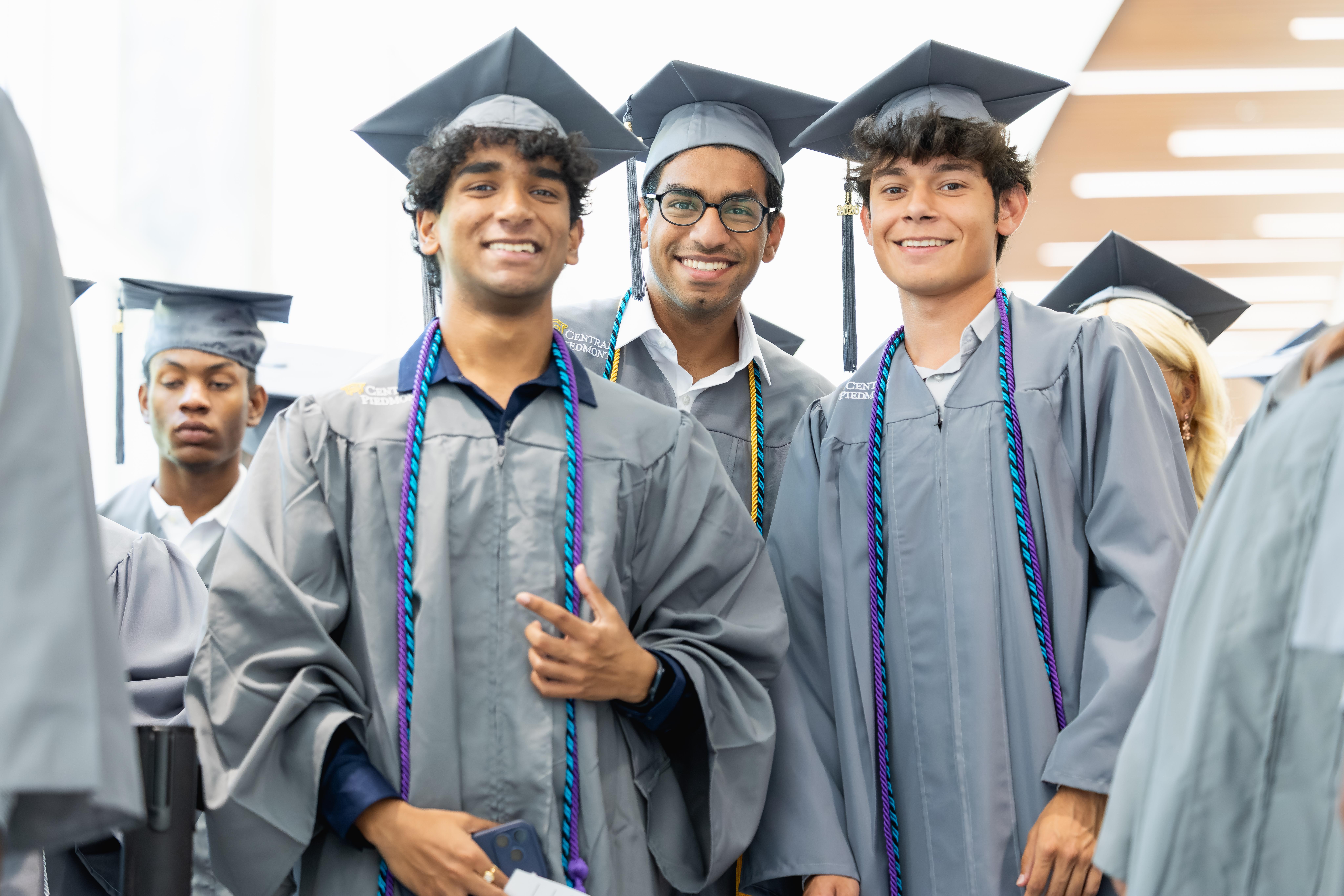 Three graduates in gray caps and gowns smile for the camera while wearing colorful stoles, with other graduates visible in the background.