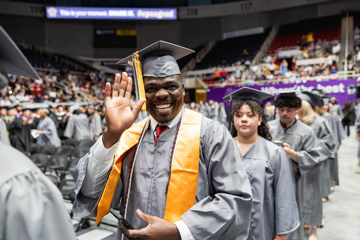 A Central Piedmont student in cap and gown, smiling and waving at the camera