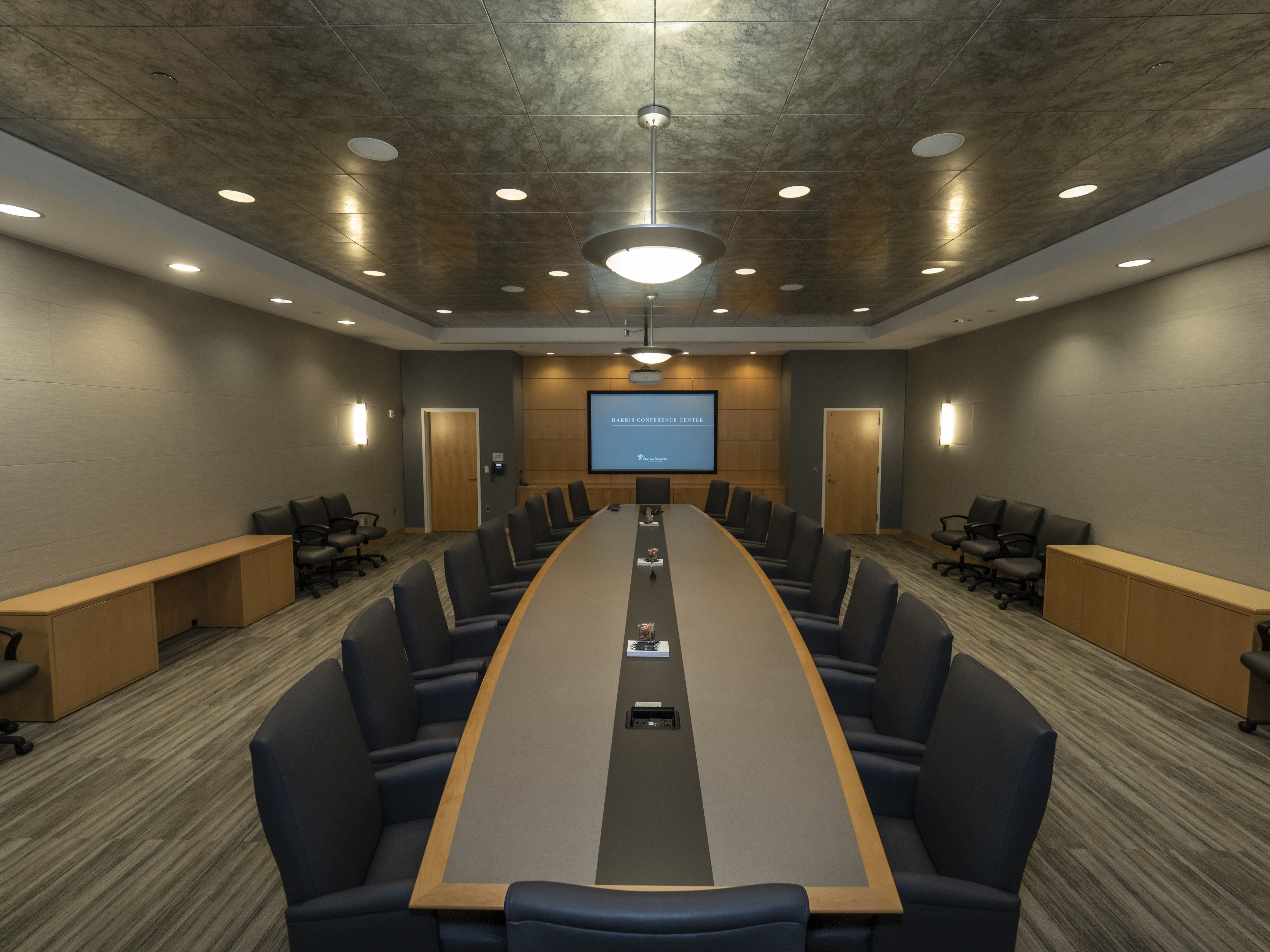 Empty conference room with long boardroom table with dark colored chairs, dramatic lighting and digital screen at the far end of the room