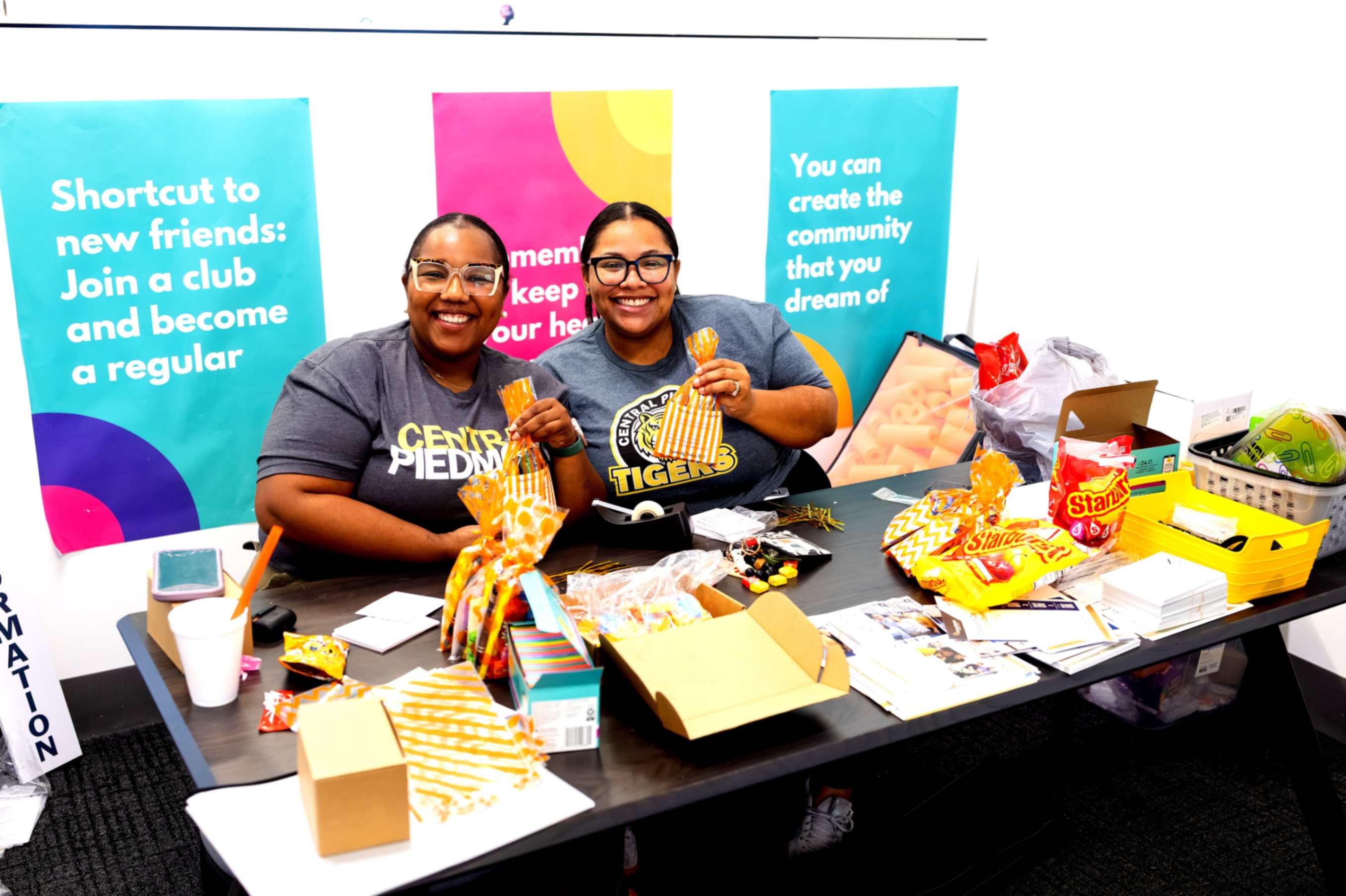 Two staff members wearing Central Piedmont shirts behind a table with papers, snacks and gift bags. Behind them are banners saying "Join a club" and "You can create the community that you dream of".