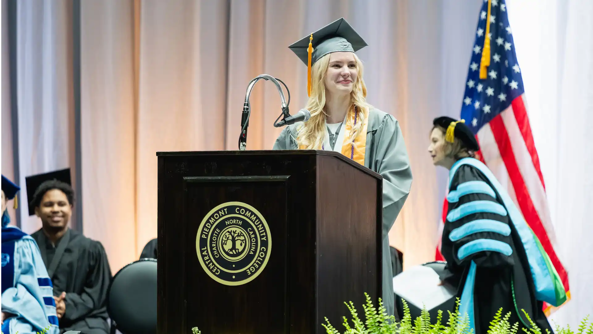 A graduate in academic regalia stands at a podium with the emblem of Piedmont Community College, addressing an audience during a commencement ceremony.