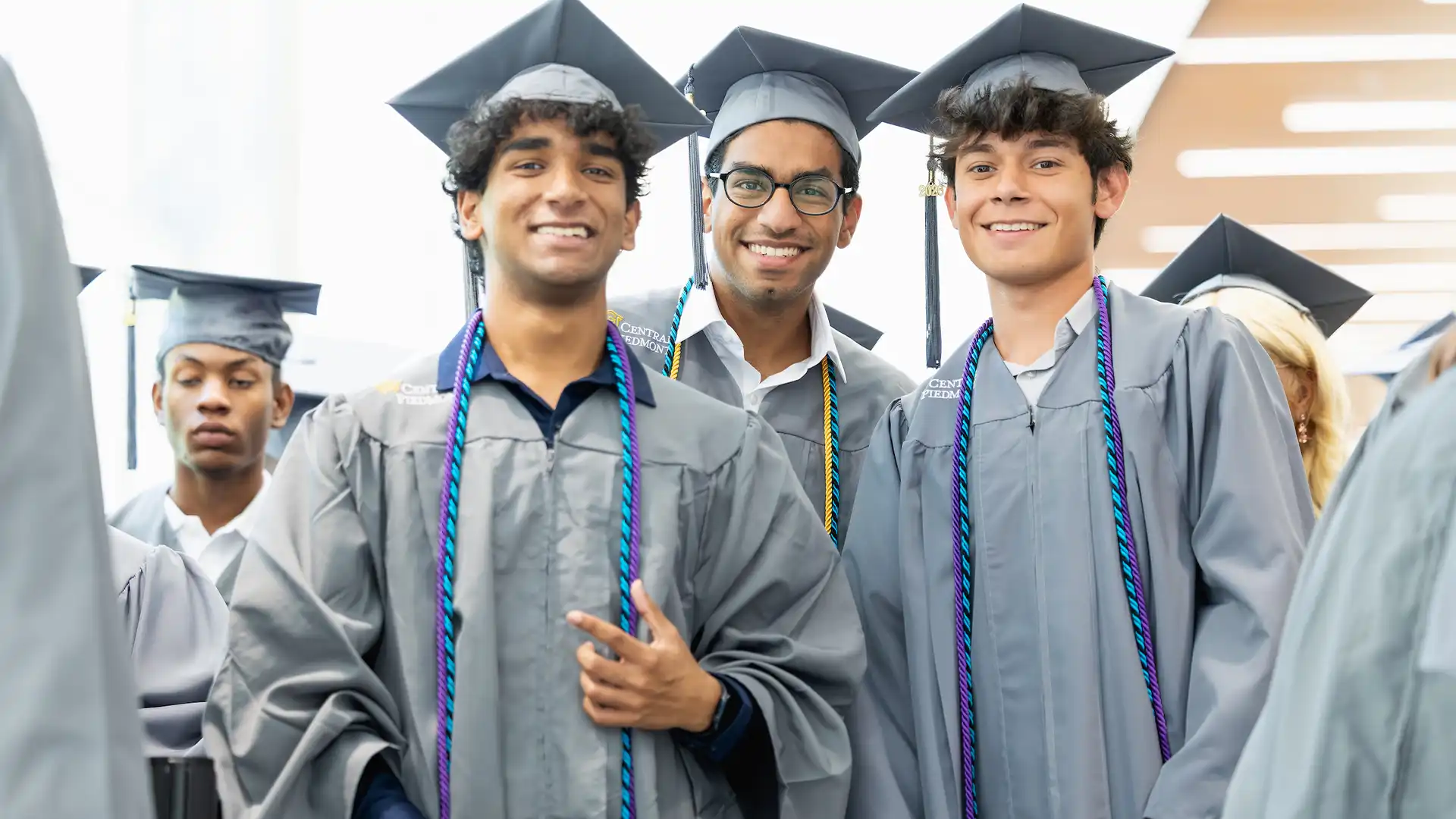 Three graduates in gray caps and gowns smile for the camera while wearing colorful stoles, with other graduates visible in the background.