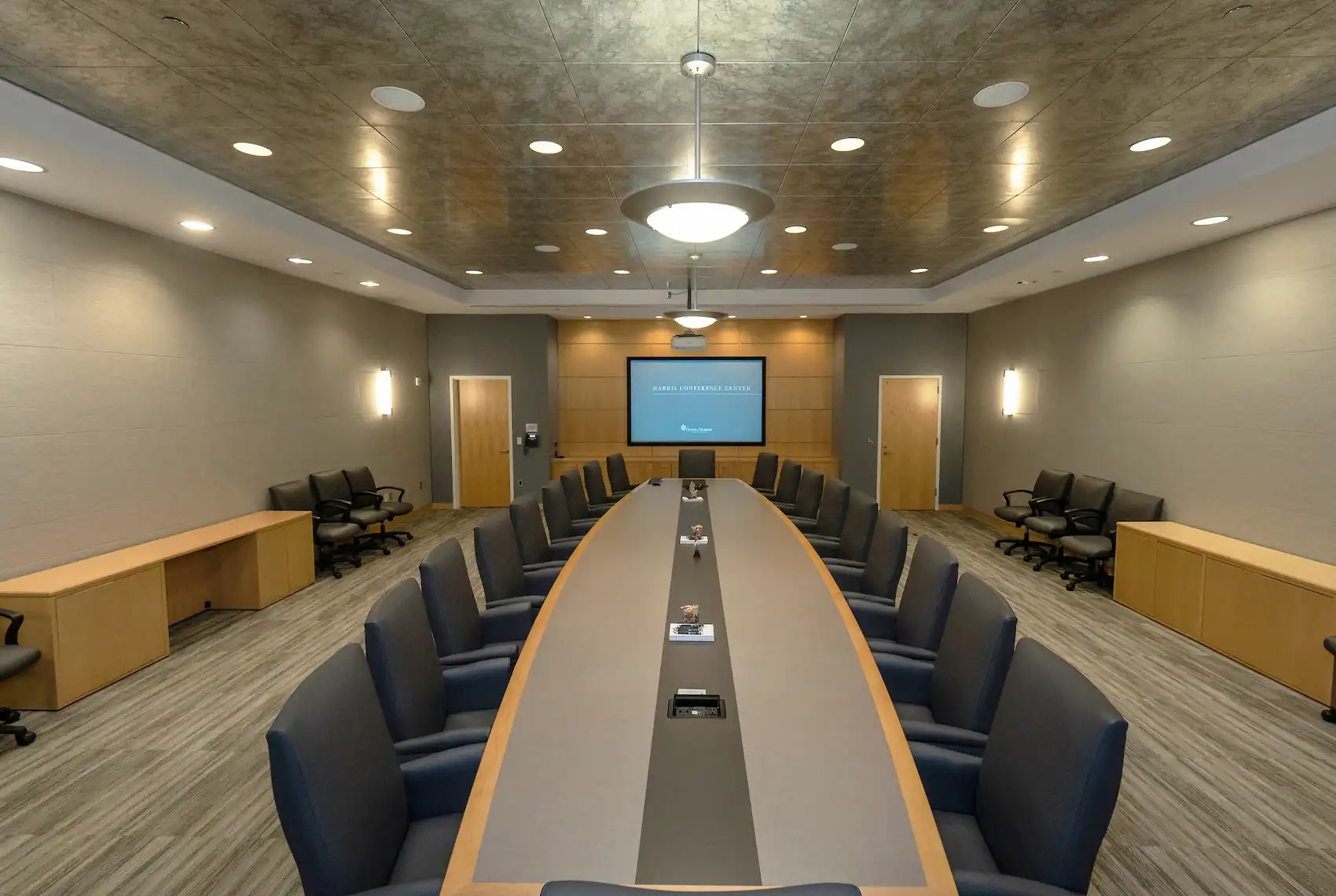 Empty conference room with long boardroom table with dark colored chairs, dramatic lighting and digital screen at the far end of the room