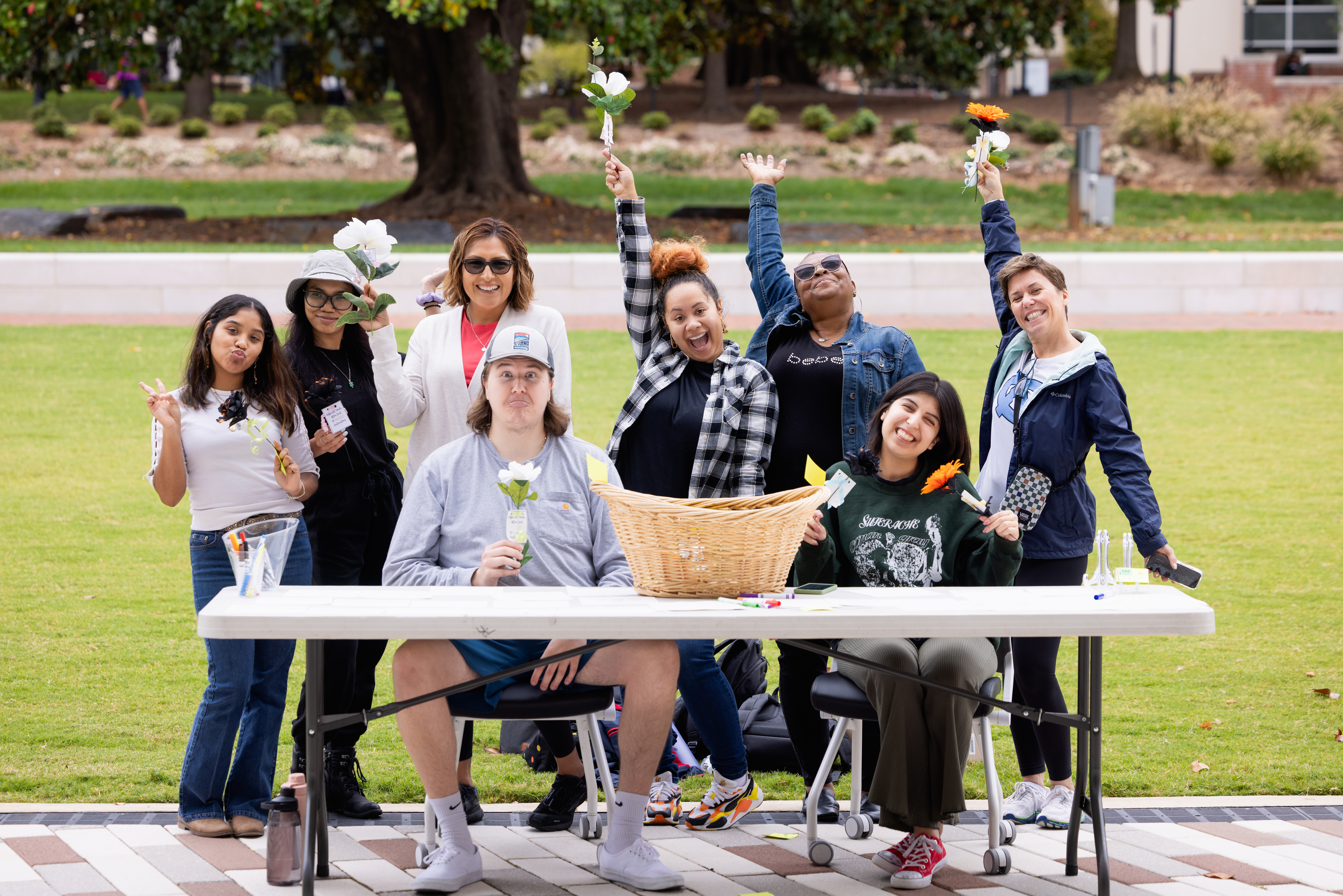 Eight students on Central campus quad where two are seated at a table and the other standing behind holding bouquets of flowers with their arms in the air.