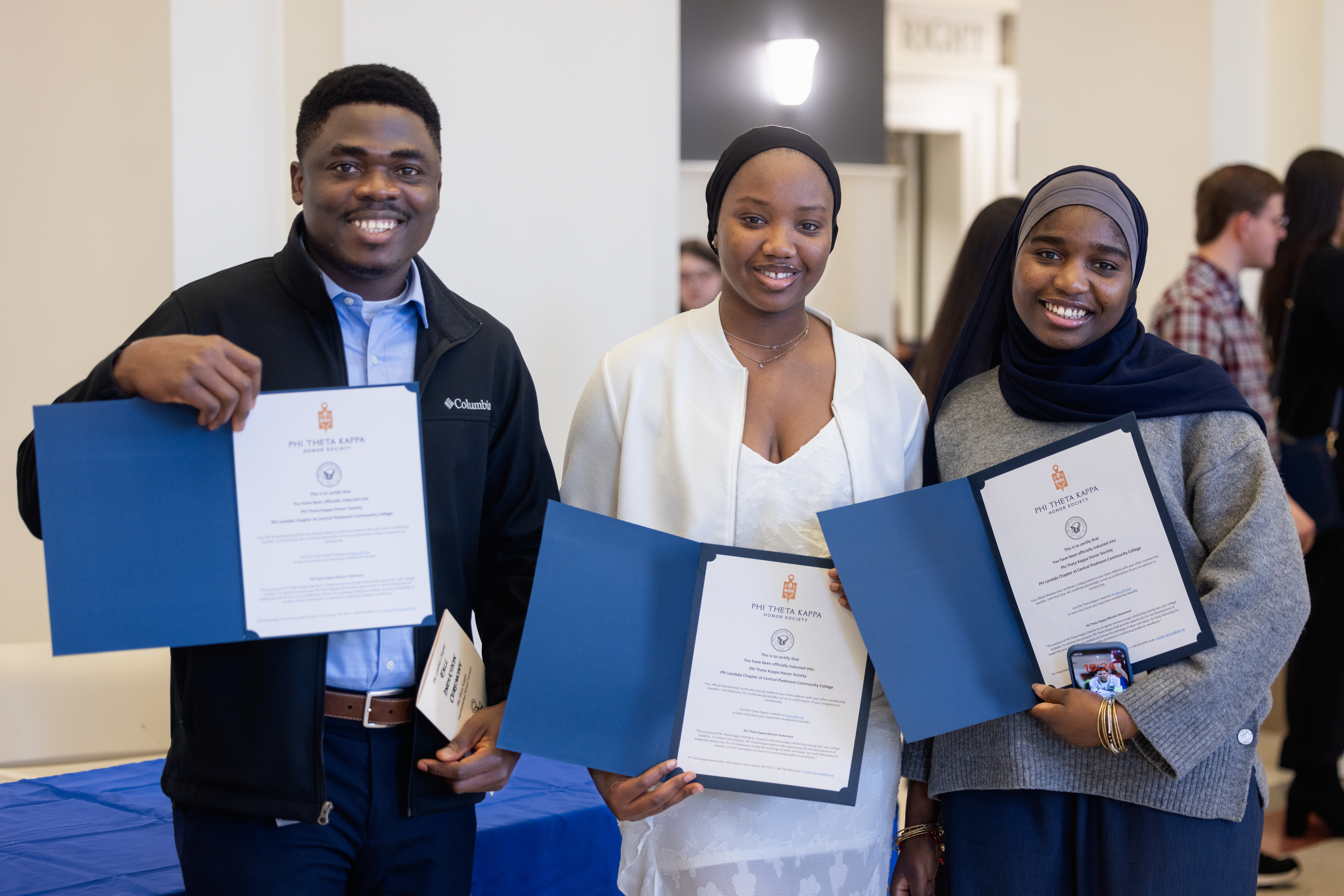 Three smiling students proudly holding up Phi Theta Kappa certificates in blue folders