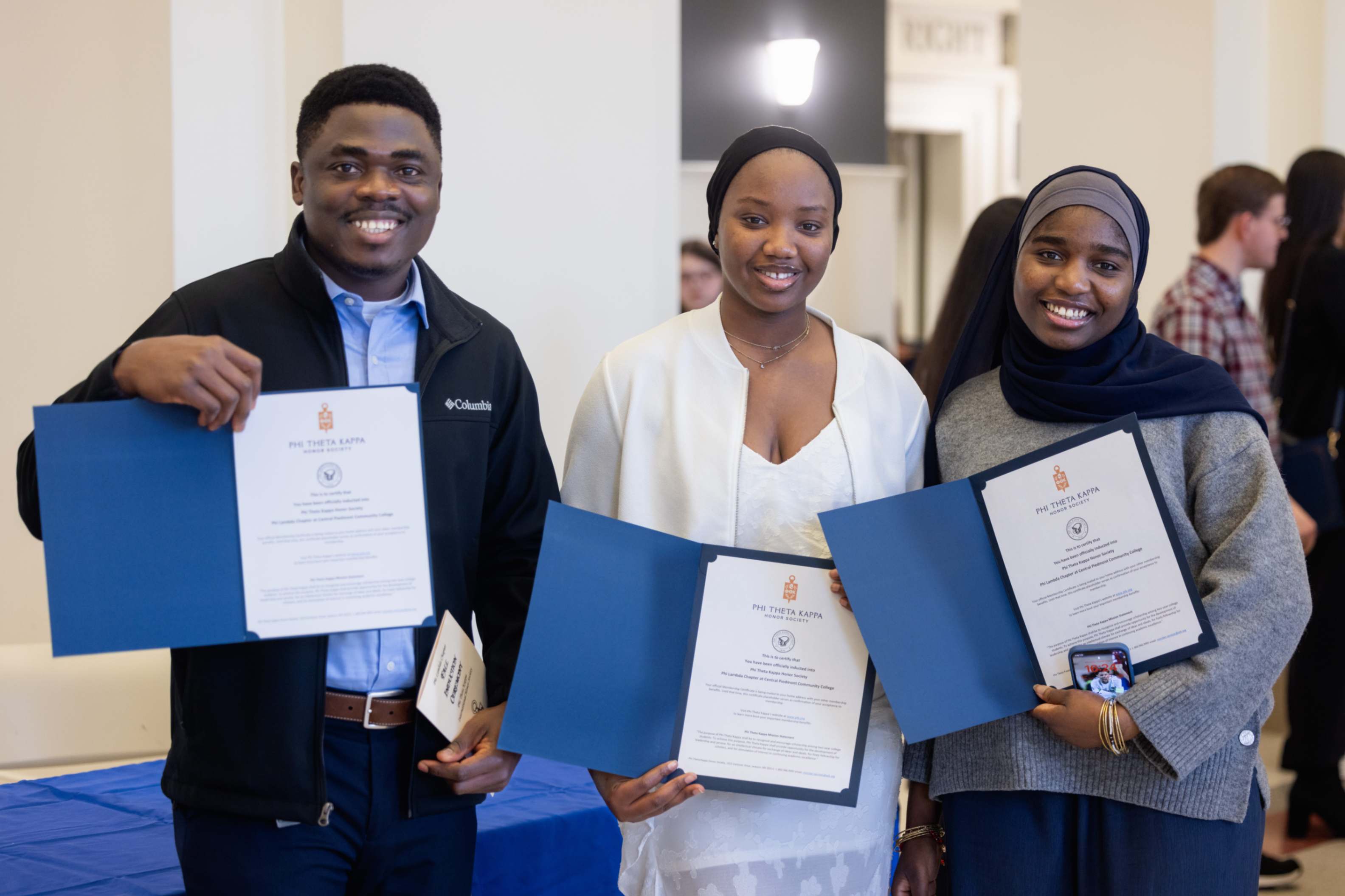 Three smiling students proudly holding up Phi Theta Kappa certificates in blue folders