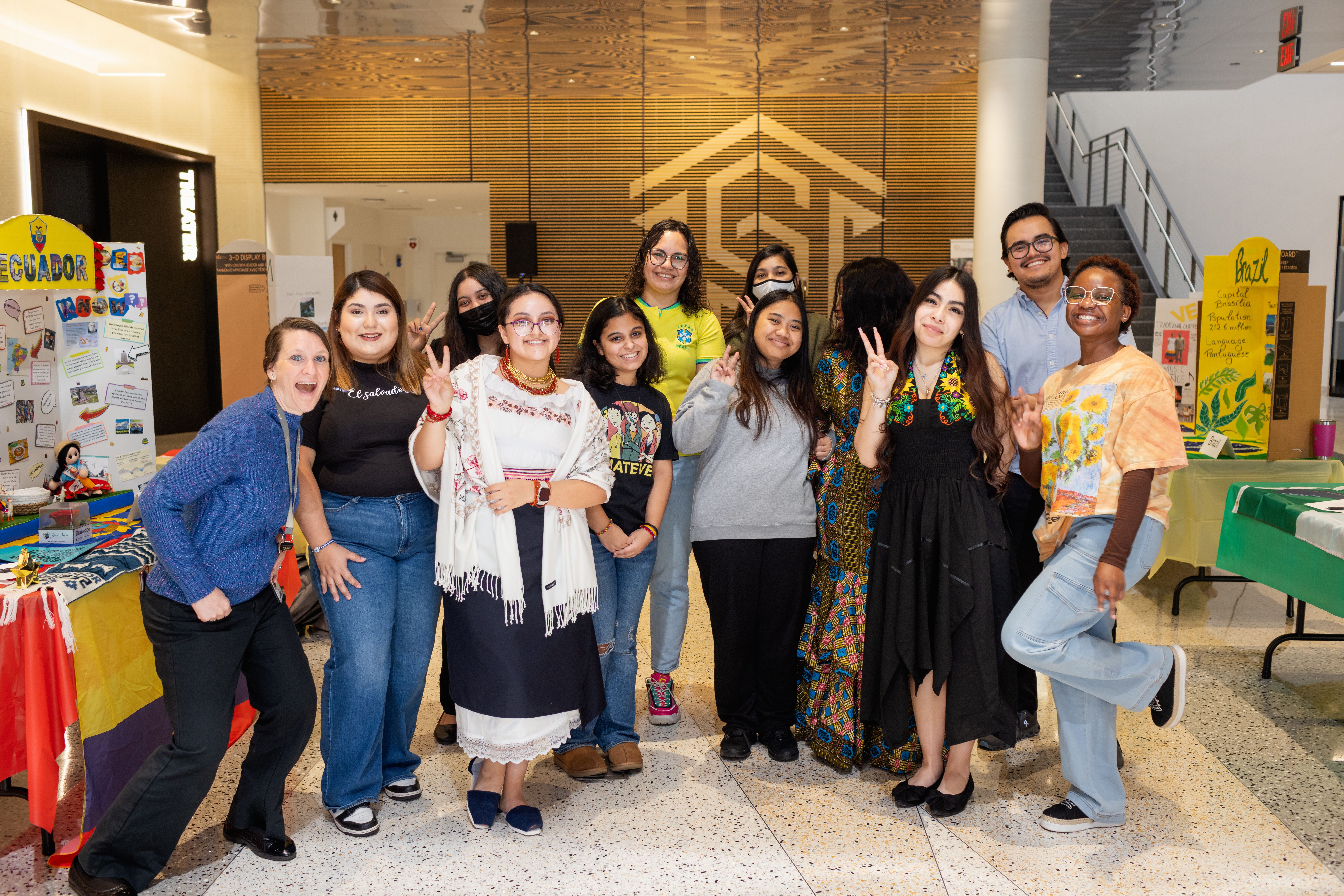 Twelve international club students outside of Parr theatre with display tables on left and right sides showing information about different countries and a large Central Piedmont logo in the background