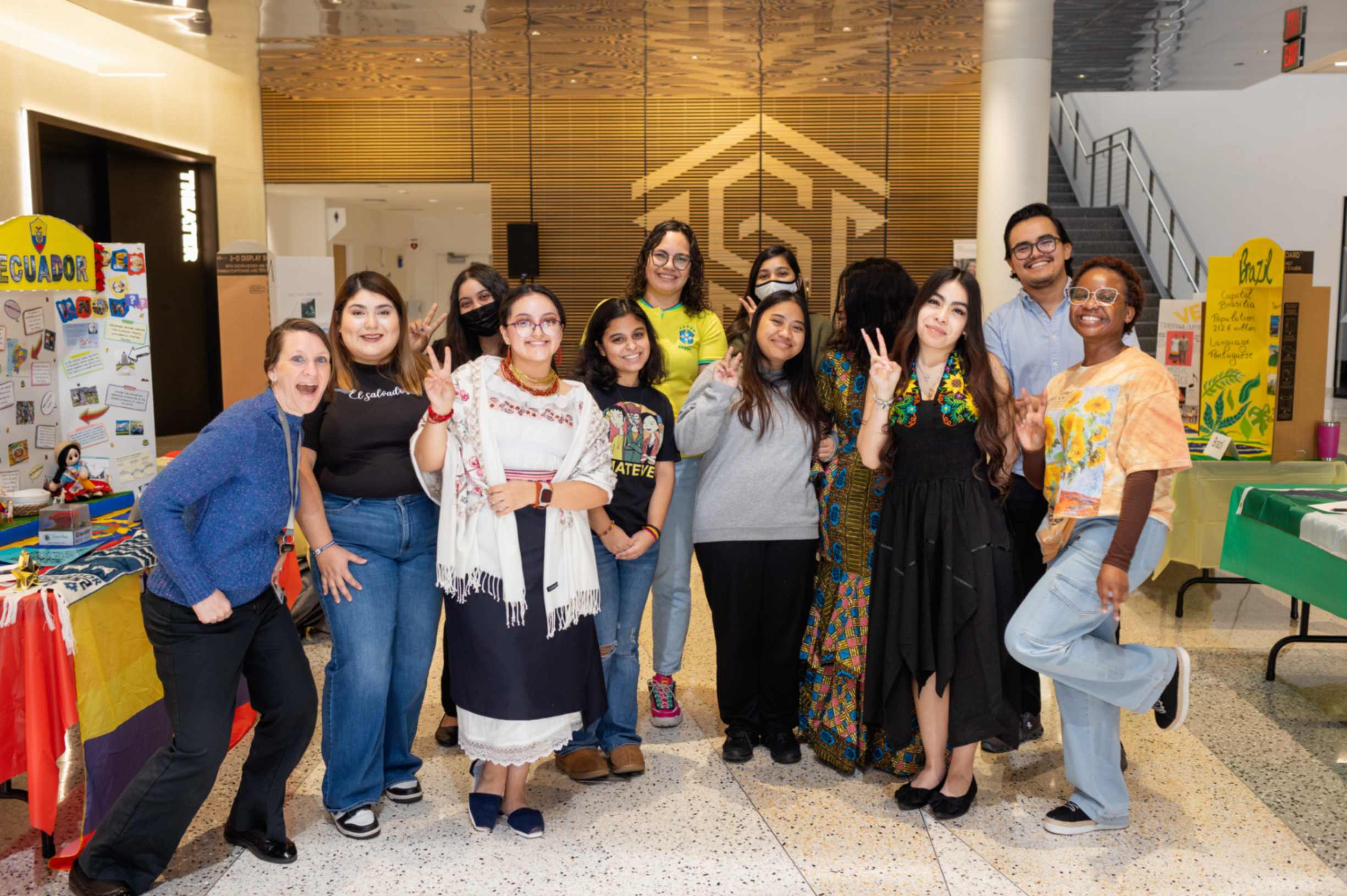 Twelve international club students outside of Parr theatre with display tables on left and right sides showing information about different countries and a large Central Piedmont logo in the background