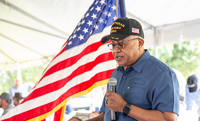 A veteran in a service cap speaking at a microphone beside a large American flag