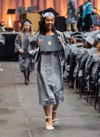 Central Piedmont student in graduation cap and gown walking down aisle excitedly showing diploma with big smile.