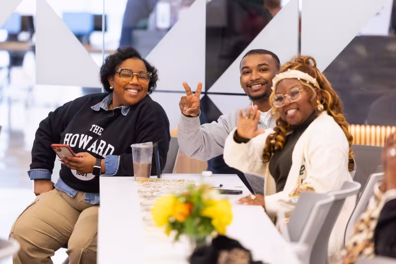 Three Central Piedmont students sitting at end of long white table inside, smiling and waving at camera.