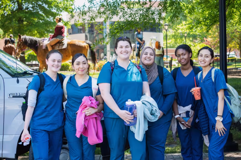 Six Central Piedmont students wearing blue scrubs and smiling for camera while students ride horses in background.