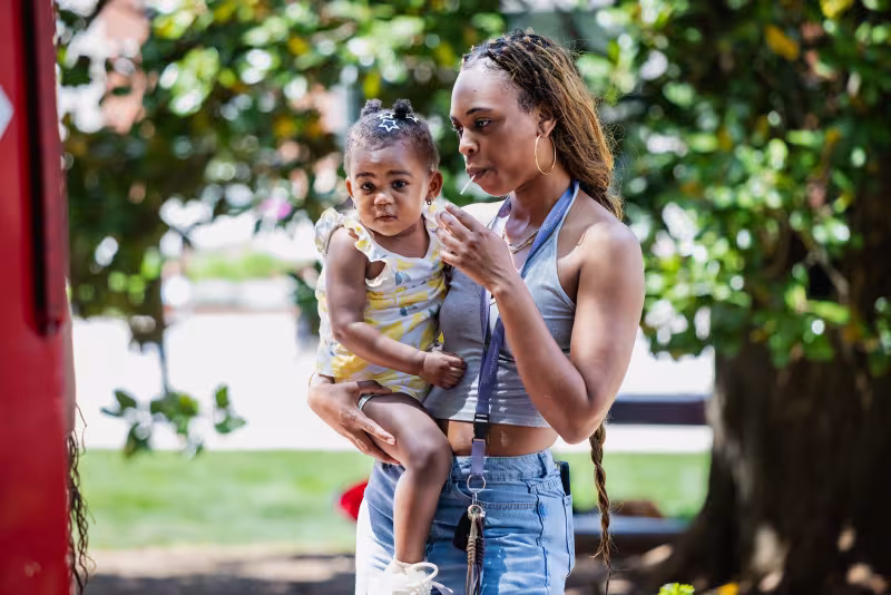 Central Piedmont student eating lollipop while holding toddler in arms outside on campus with trees in background.