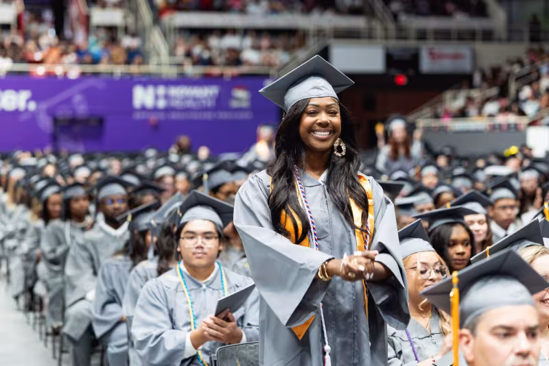 Graduate standing and smiling among seated classmates at commencement