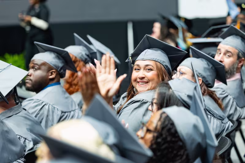 Graduate wearing cap and gown waving and smiling at the camera during commencement ceremony