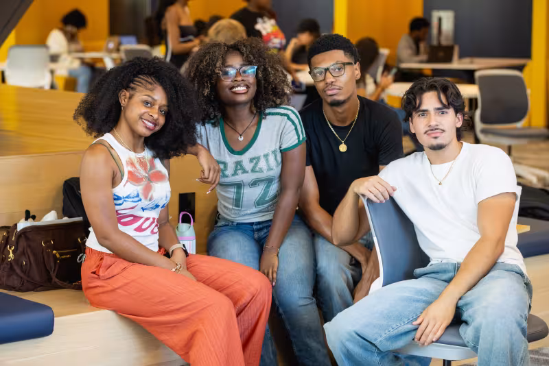 Group of four students in the library on Central Campus looking at the camera and smiling.