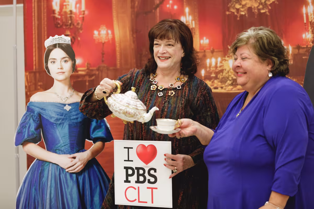 Two WTVI PBS Charlotte supporters pouring tea beside a Queen Victoria cutout