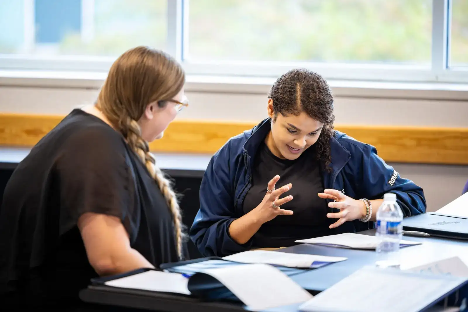 Two students discussing notes across a table in a classroom