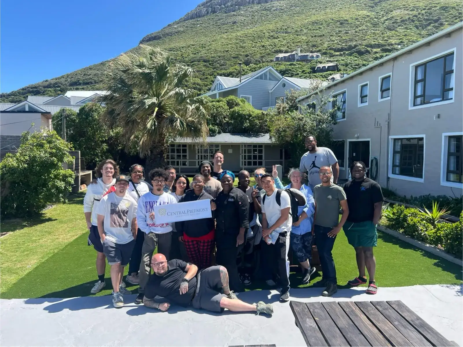 Group of study abroad students in tropical country holding “Central Piedmont” sign with housing in background.