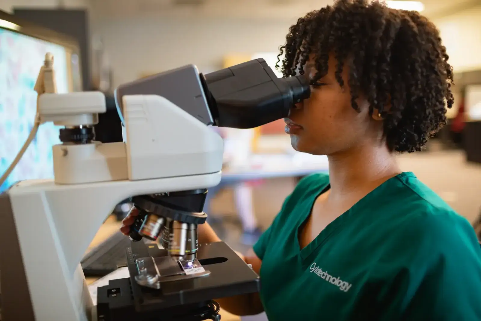 Central Piedmont student wearing cytotechnology scrubs looking through microscope in lab.