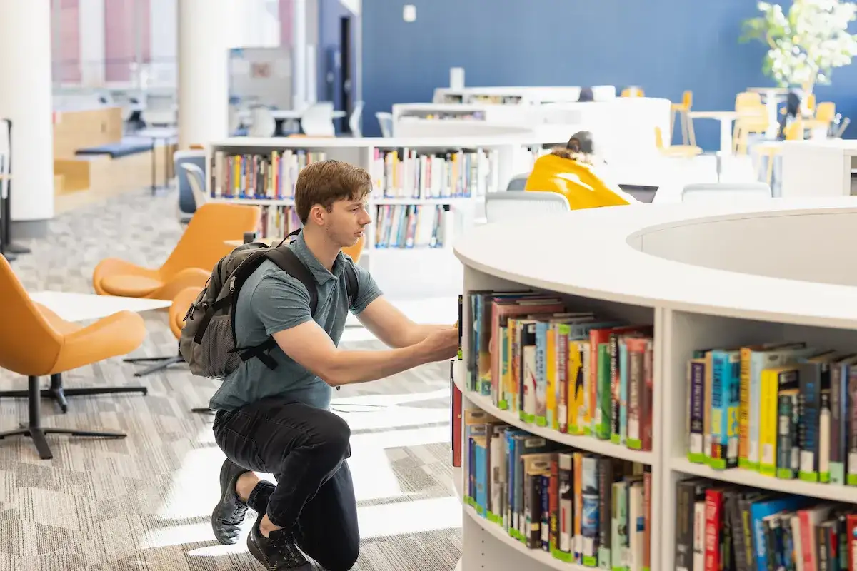 Student kneeling to look at books on the library shelf