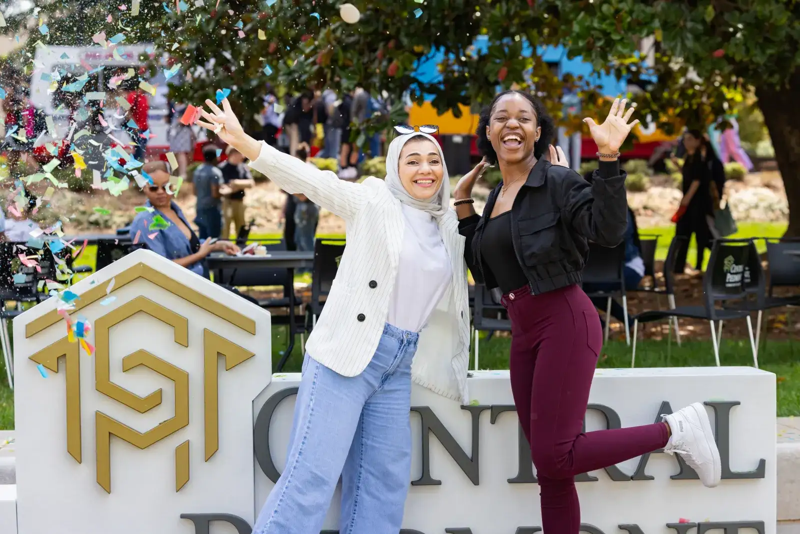 Two students celebrating and posing for camera in front of large Central Piedmont signage with confetti all around.
