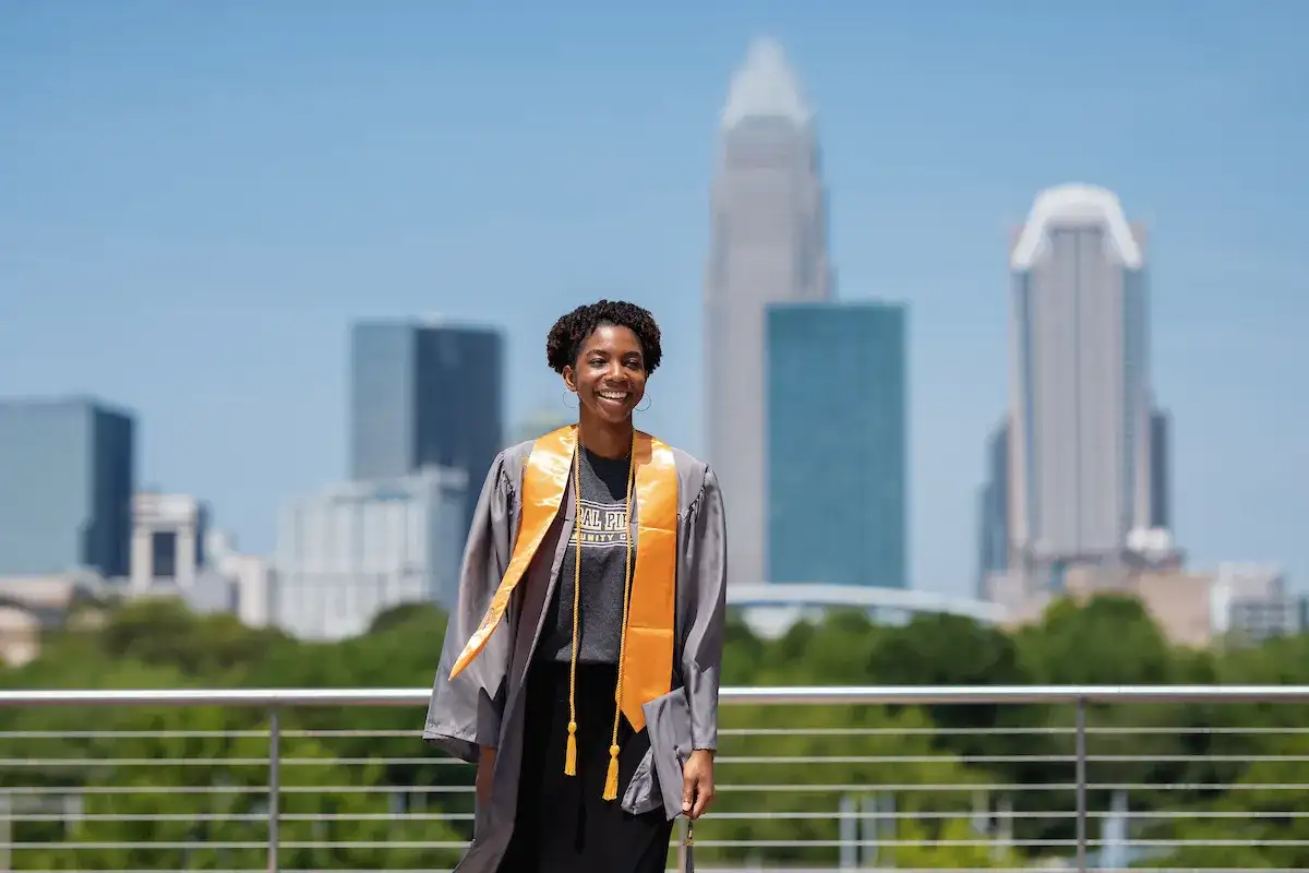 Student in graduation robe and tassels standing on the student center balcony with the Charlotte skyline in the background