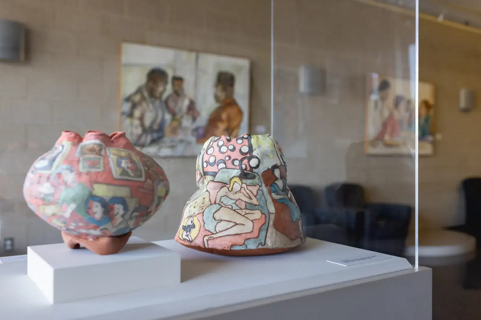 Two multi-colored pottery pieces in glass case with artwork hanging on the wall in the background