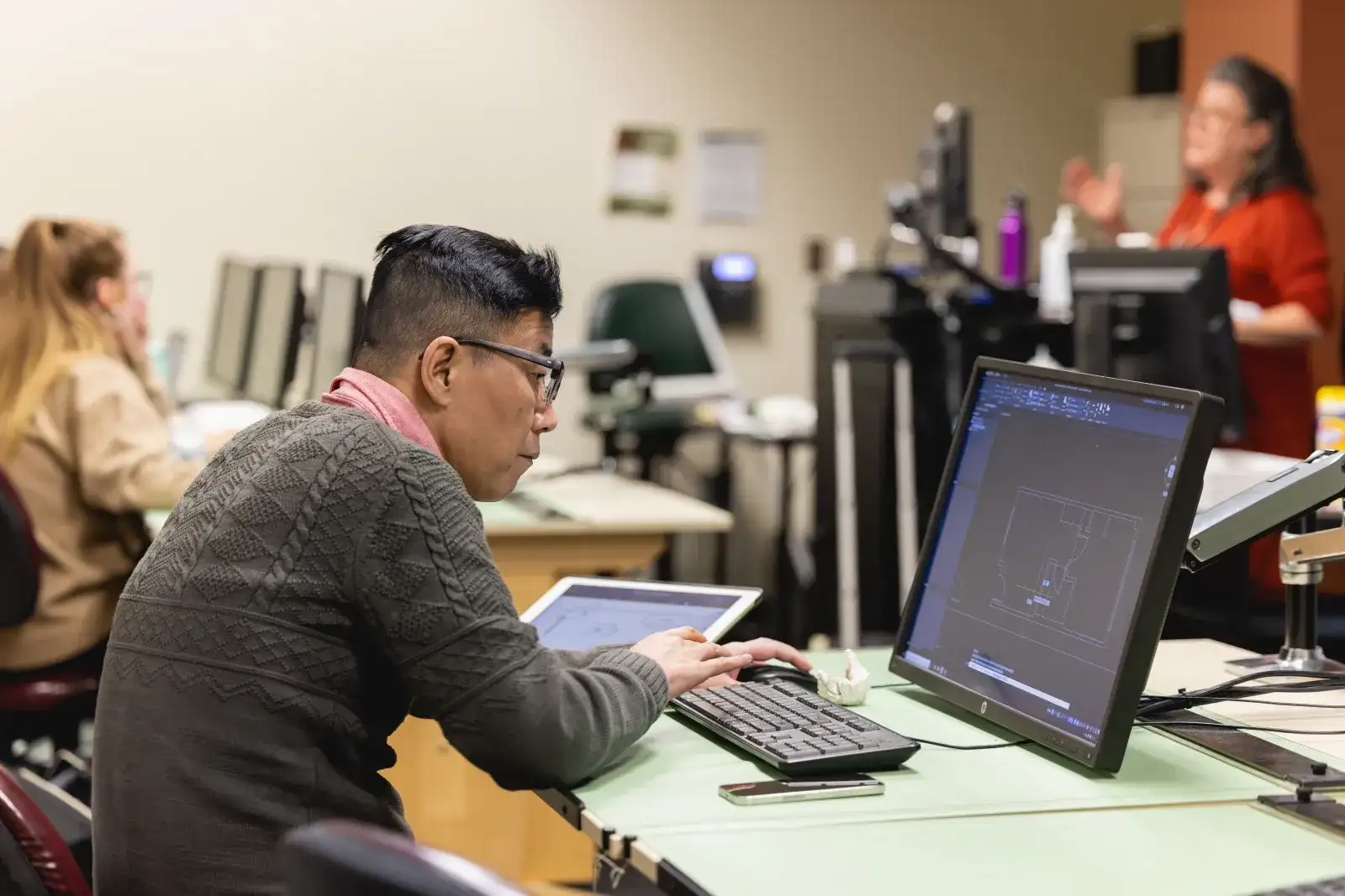 Student looking intently at a computer screen with another student and instructor in the background