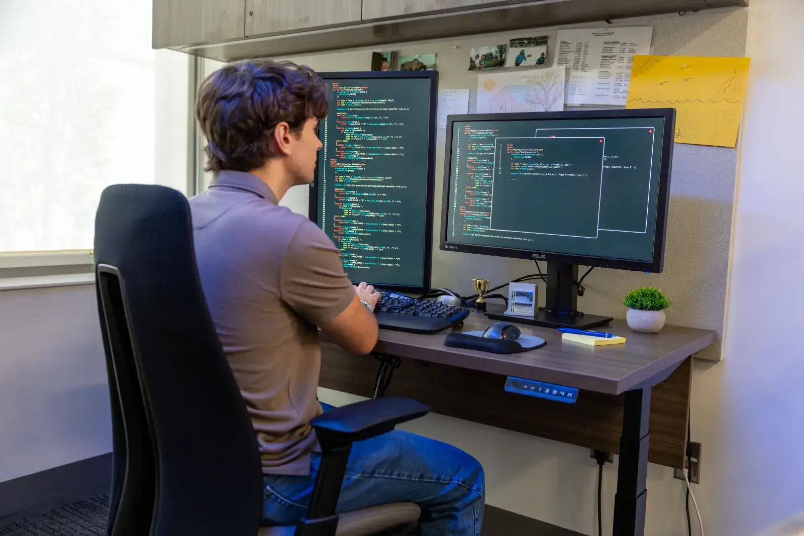 Student with his back to the camera sitting at a desk with two large monitors both with overlapping screens of code data written in green text