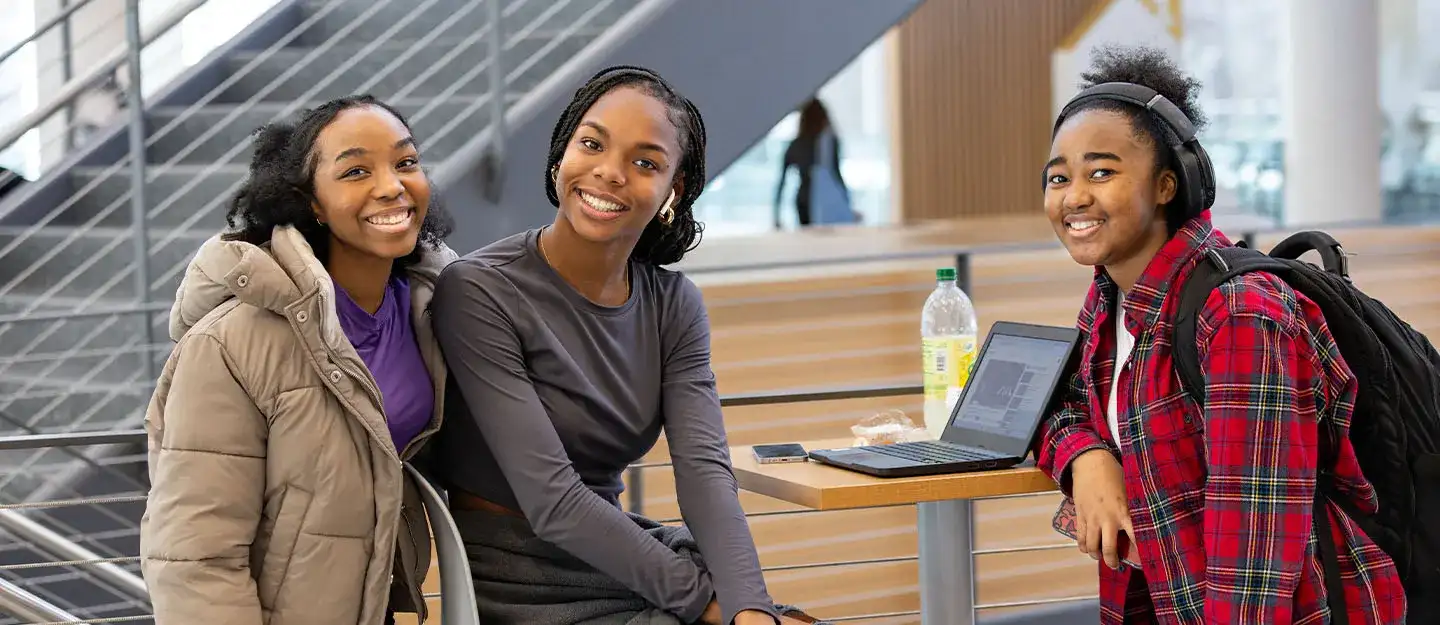 Three students smiling together at a table with a laptop in a campus common area