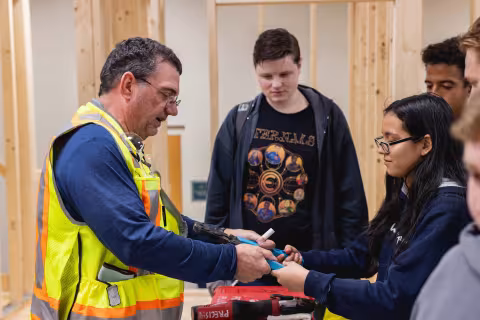 Instructor in a high-visibility vest demonstrating a power tool to construction students